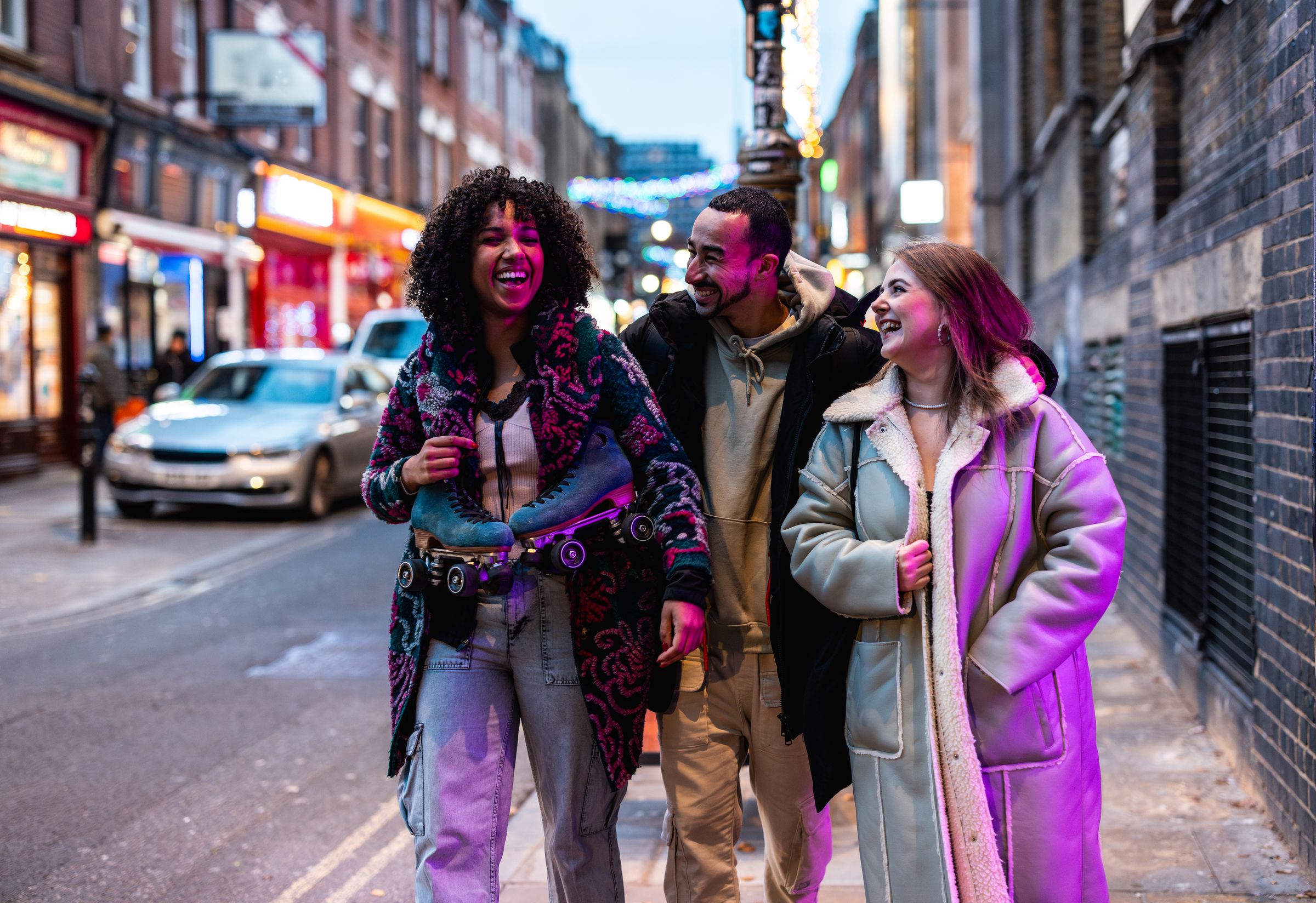 Three friends laughing on a city street at dusk, one carrying roller skates